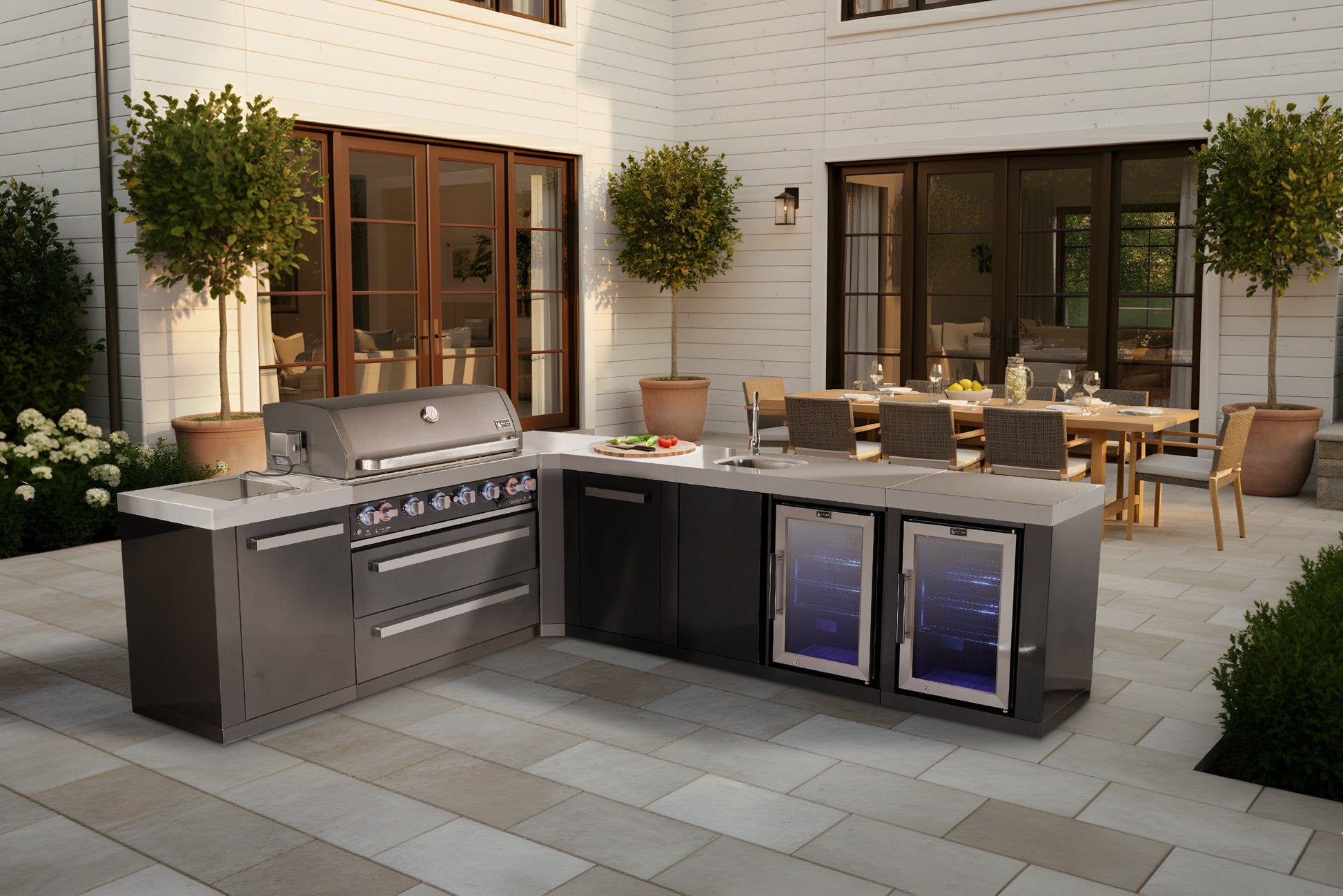 Mont Alpi outdoor kitchen with stainless grill, burners, fridge, counter space, dining table, and lush plants nearby.