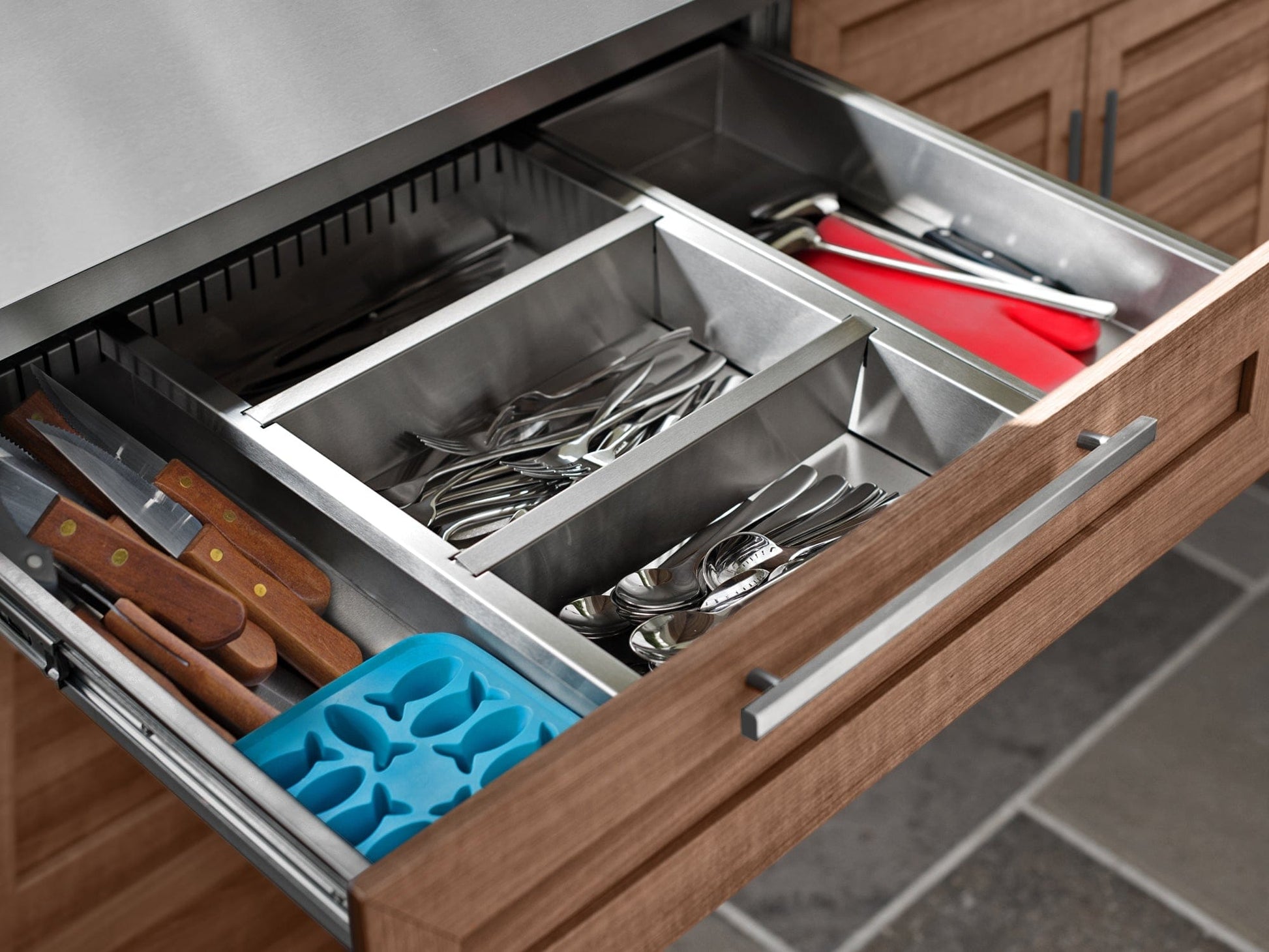 Sleek outdoor kitchen drawer with organized cutlery, red cutting board, and stainless steel cabinets nearby.