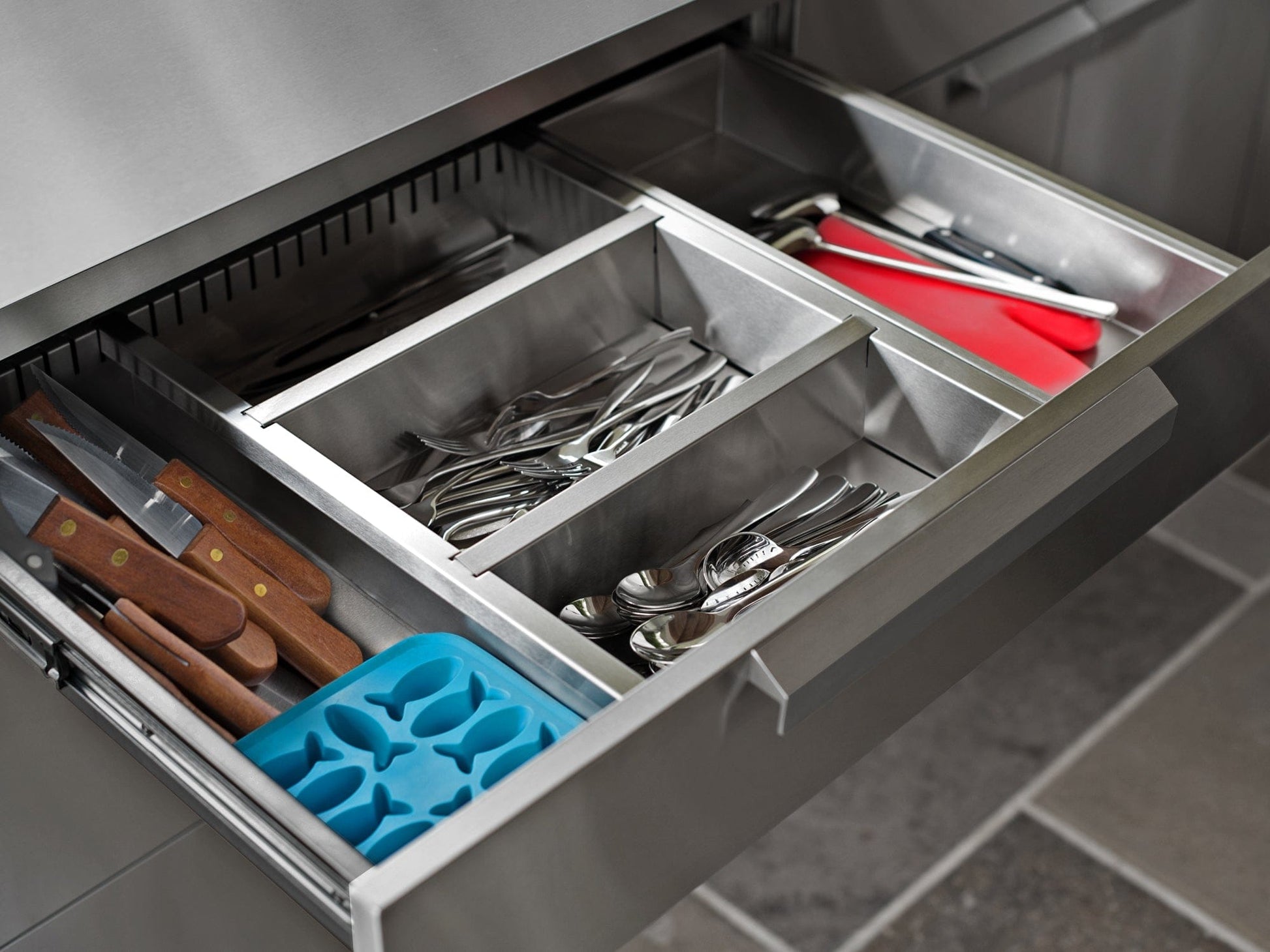 Organized kitchen drawer with utensils, knives, and blue ice tray in a premium outdoor stainless steel kitchen setup.
