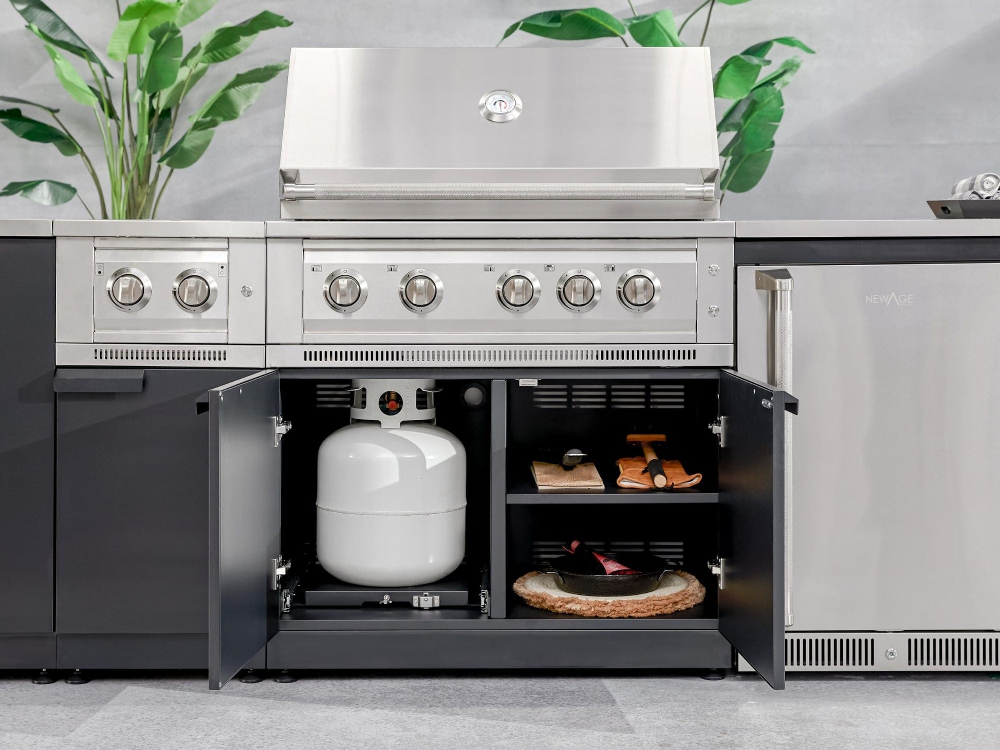 Stainless steel outdoor grill with open cabinet showing propane tank, grill tools, and pizza stone storage.