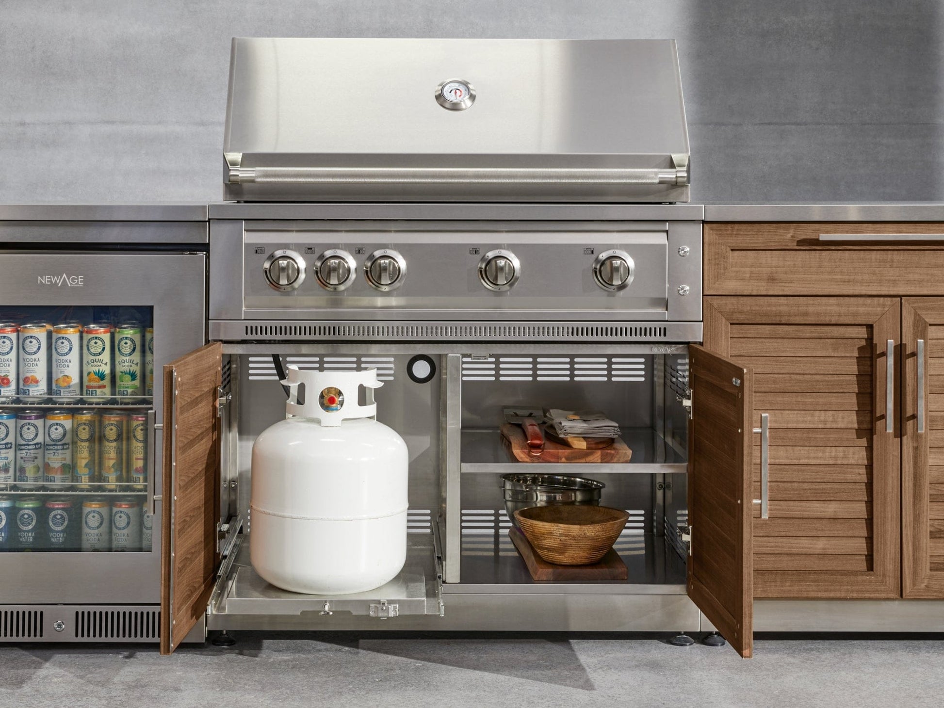 Outdoor stainless steel grill with open doors showing storage, propane tank, and kitchen accessories.