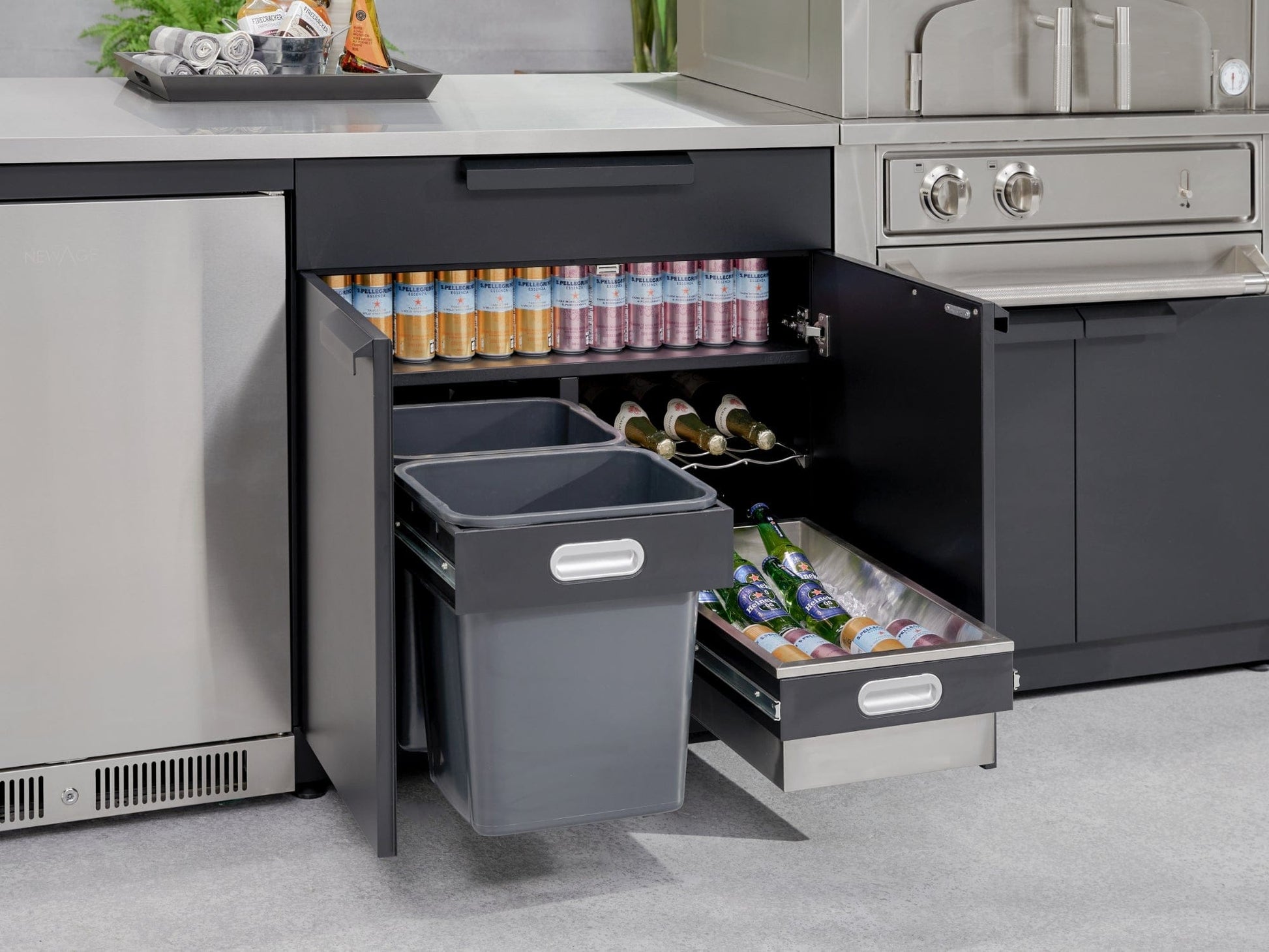 Outdoor kitchen cabinets with open drawers showing drinks, bottles, cans, and two trash bins in a sleek finish.