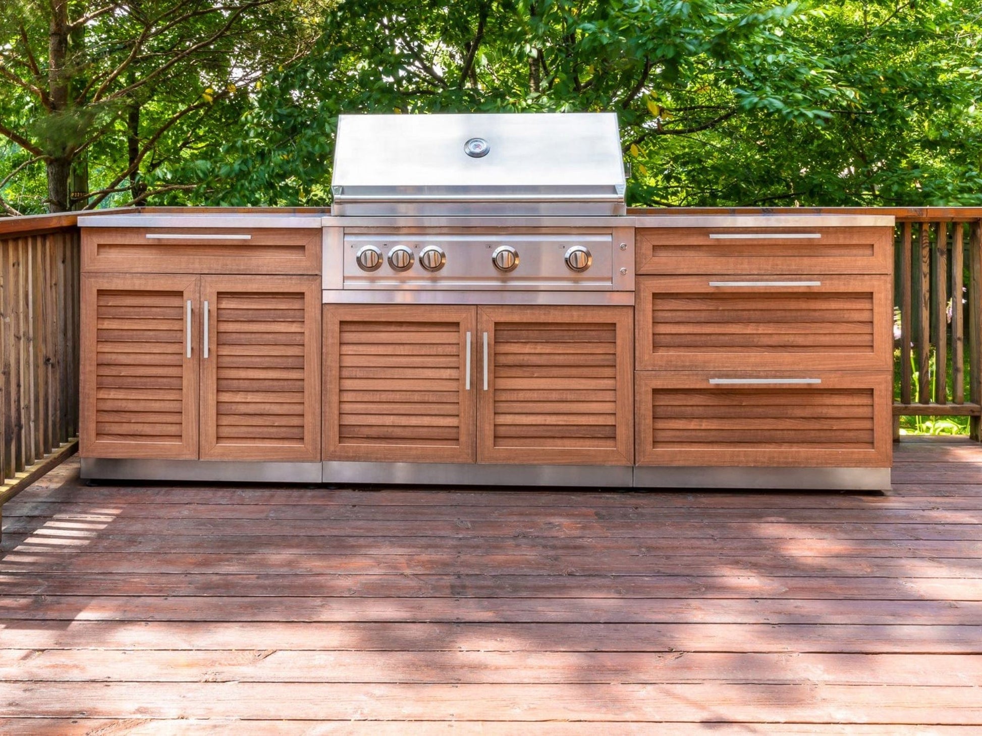 Premium outdoor kitchen grill with modular cabinets, sink, and fridge on wood deck amid lush trees.