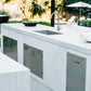 Premium outdoor kitchen with sink, cabinets, 2-bin trash drawer, and lush garden backdrop for luxury entertaining.