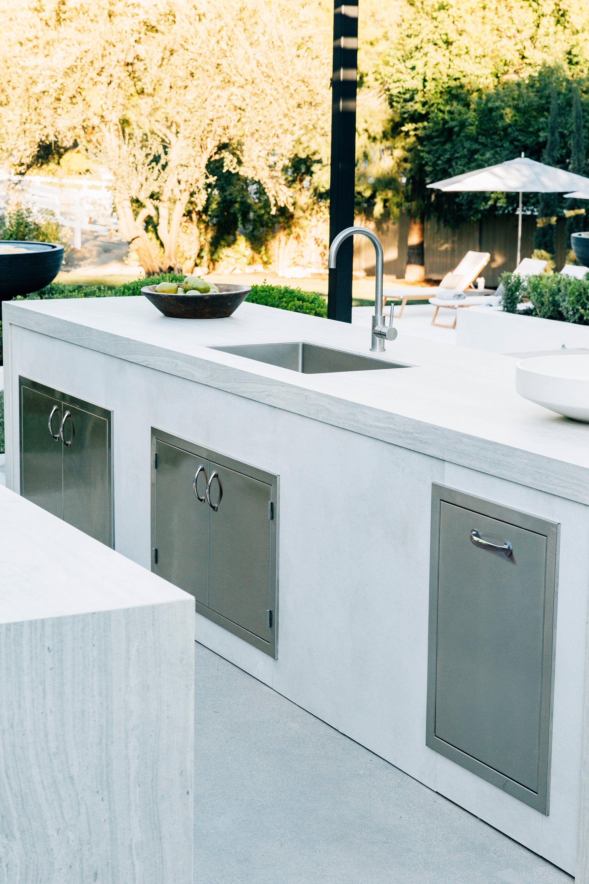 Premium outdoor kitchen with sink, cabinets, 2-bin trash drawer, and lush garden backdrop for luxury entertaining.