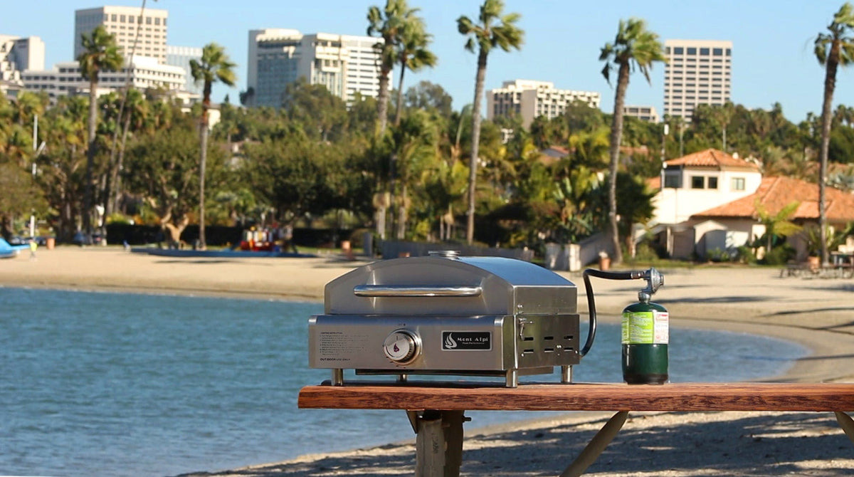 Mont Alpi Pizza Ovens Mont Alpi 3-in-1 Portable Grill, Griddle, and Pizza Oven / Stainless Steel / MA-3N1 sitting on a picnic table by the beach